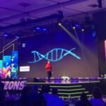 Engel Fonseca en el escenario de EY Horizons 2026 frente a una pantalla con una hélice de ADN y el logo de Curioxoma