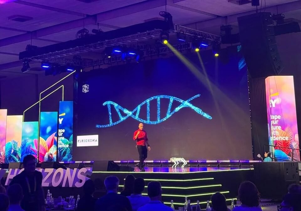Engel Fonseca en el escenario de EY Horizons 2026 frente a una pantalla con una hélice de ADN y el logo de Curioxoma