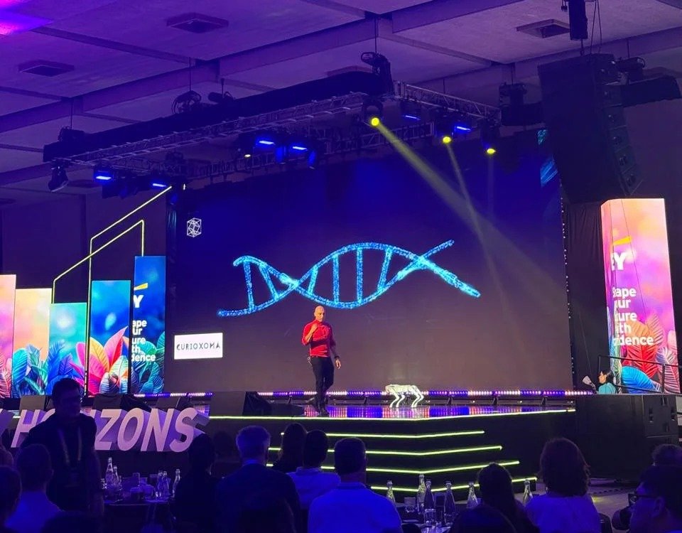 Engel Fonseca en el escenario de EY Horizons 2026 frente a una pantalla con una hélice de ADN y el logo de Curioxoma