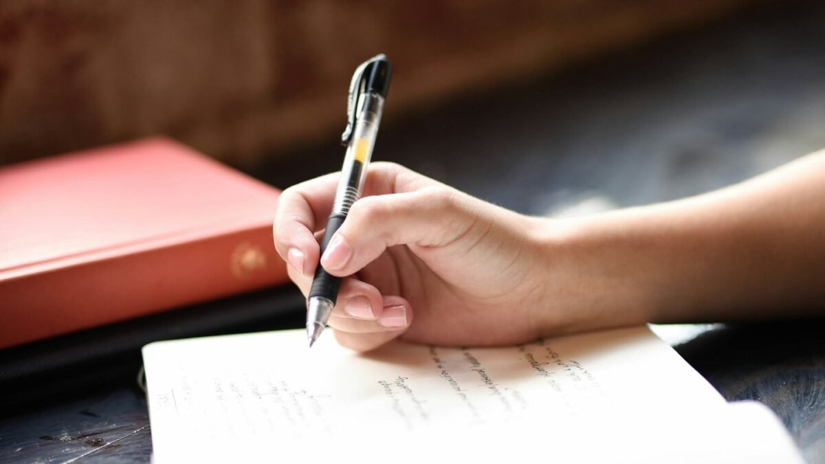 Mano escribiendo con pluma en un cuaderno junto a una ventana