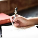 Mano escribiendo con pluma en un cuaderno junto a una ventana